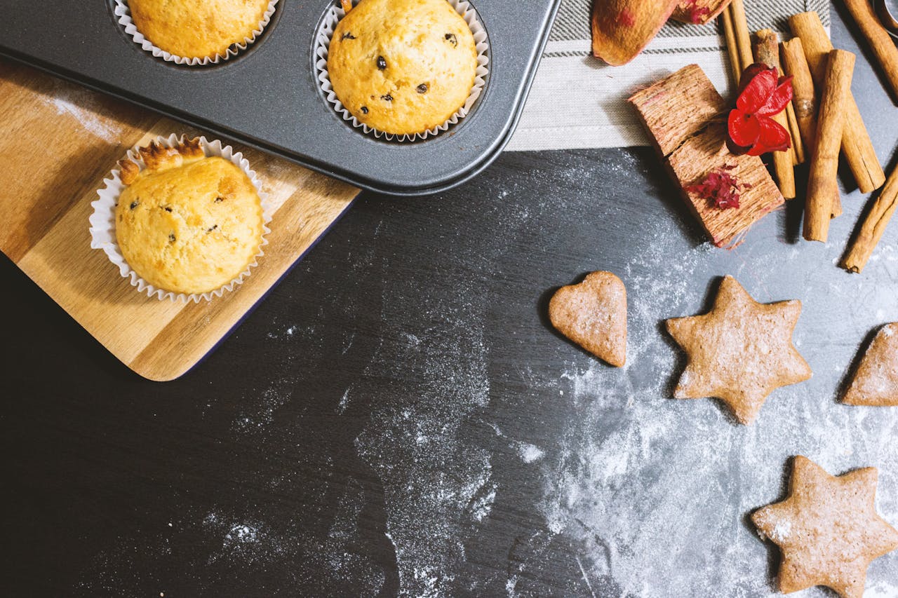 hero-img A delicious arrangement of muffins and cookies with cinnamon and flour on a black countertop.